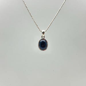 Puede incluir: Un collar de plata con un colgante ovalado azul oscuro. El colgante tiene un borde texturizado de color plateado. La cadena es un dise&ntilde;o de cuerda fina y retorcida. El fondo es gris claro liso.