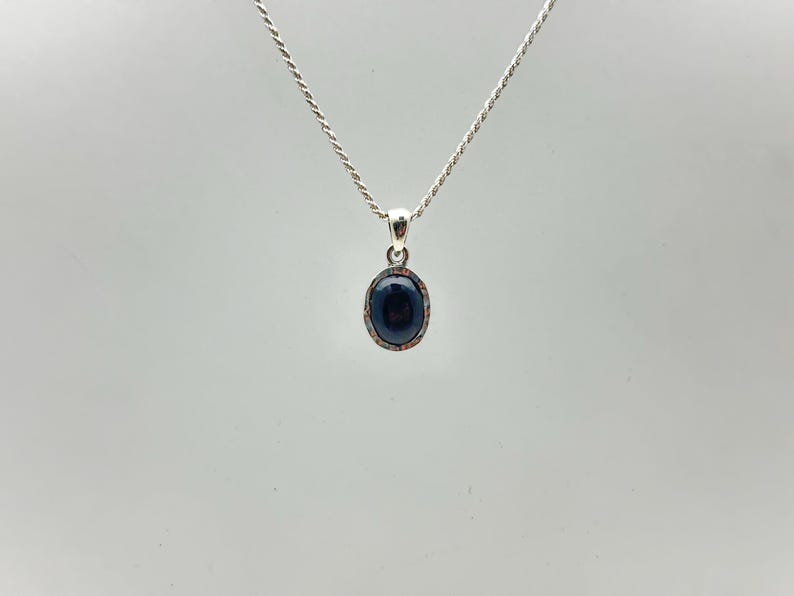 Puede incluir: Un collar de plata con un colgante ovalado azul oscuro. El colgante tiene un borde texturizado de color plateado. La cadena es un dise&ntilde;o de cuerda fina y retorcida. El fondo es gris claro liso.