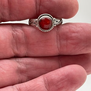 May include: A silver ring with a round, faceted, red gemstone. The ring has a twisted rope design around the gemstone and decorative swirls on the sides. The ring is held in a person's hand.