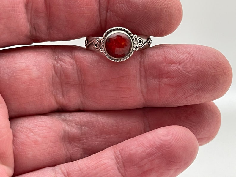 May include: A silver ring with a round, faceted, red gemstone. The ring has a twisted rope design around the gemstone and decorative swirls on the sides. The ring is held in a person's hand.