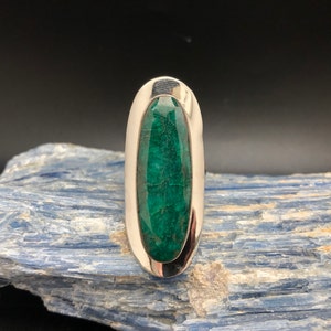 May include: A silver ring with a large, oval, deep green stone. The stone is set in a polished silver band. The ring is displayed on a textured blue surface against a dark background. The ring's design is simple yet elegant.