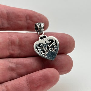 Puede incluir: Un colgante de plata en forma de coraz&oacute;n con intrincados detalles de filigrana. El colgante tiene una anilla en la parte superior para sujetarlo a una cadena.