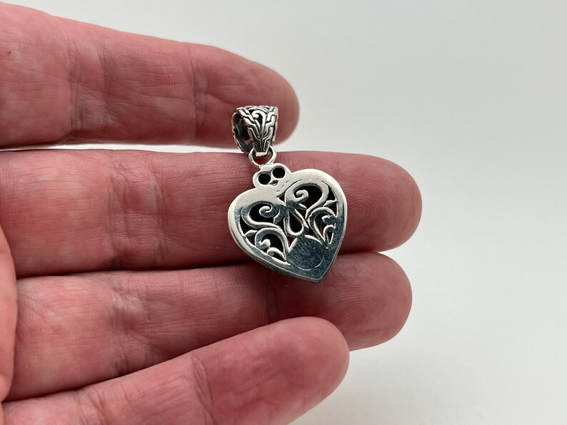 Puede incluir: Un colgante de plata en forma de coraz&oacute;n con intrincados detalles de filigrana. El colgante tiene una anilla en la parte superior para sujetarlo a una cadena.