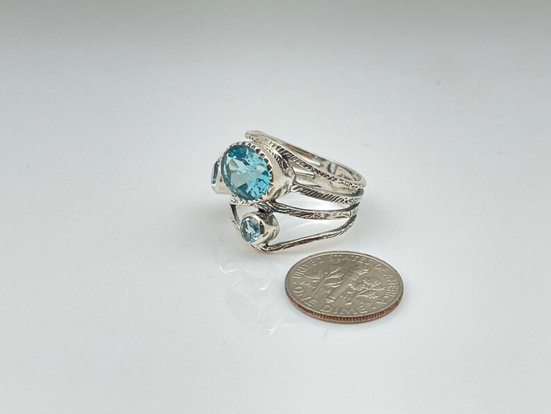 May include: A silver ring with three blue topaz gemstones. The ring has a textured band and is set with a larger oval-shaped stone and two smaller round stones.