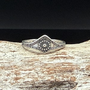 May include: A silver ring with an ornate design. The ring features a central diamond shape with a sunburst pattern and dotted accents. The band is also decorated with small dots. The ring is displayed on a piece of wood.