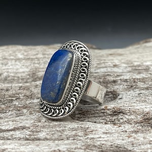 May include: A silver ring with a large, rectangular lapis lazuli stone set in a decorative silver bezel. The ring has a detailed, ornate design.