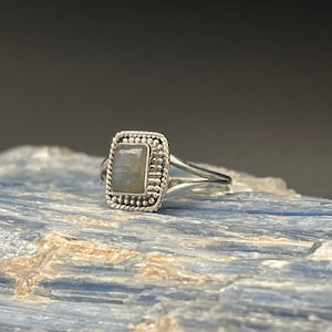 May include: A silver ring with a square-shaped labradorite gemstone set in a beaded bezel. The ring has a simple band.