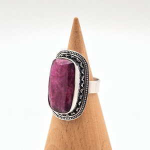 May include: A silver ring with a large, rectangular, deep pink gemstone set in a silver bezel. The ring is on a wooden display stand.