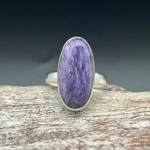 Op de afbeelding: Een zilveren ring met een ovale, paarse en witte edelsteen. De ring ligt op een houten oppervlak.