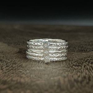 May include: A set of five silver stacking rings with different textured patterns. The rings are stacked on top of each other and are sitting on a wooden surface.