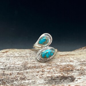 May include: A silver ring with two turquoise stones set in a unique, asymmetrical design. The stones are a light blue color with brown veins running through them.