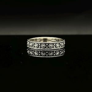 May include: A silver ring with an intricate floral pattern. The ring's design features small, circular accents and a repeating pattern around the band. The ring is displayed on a reflective black surface, enhancing its details.