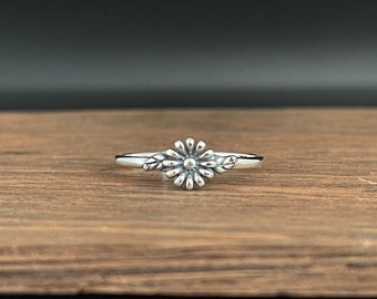 Könnte beinhalten: Ein silberner Ring mit einem Gänseblümchen-Design. Der Ring hat eine zentrale Gänseblümchenblüte mit Blütenblättern und Blättern auf beiden Seiten. Der Ring wird auf einer Holzoberfläche präsentiert.