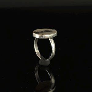 K&ouml;nnte beinhalten: Ein silberner Ring mit einem gro&szlig;en, ovalen Stein. Der Stein hat eine cremefarbene Basis mit dunkelgrauen und schwarzen Mustern. Der Ring ist vor einem schwarzen Hintergrund platziert, mit einer Reflexion darunter.