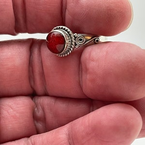 May include: A silver ring featuring a round, reddish-orange stone. The stone is set in a decorative bezel with a twisted rope design. The ring's band has a delicate, swirling pattern on the sides, adding to its ornate appeal.