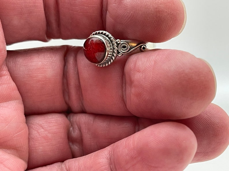 May include: A silver ring featuring a round, reddish-orange stone. The stone is set in a decorative bezel with a twisted rope design. The ring's band has a delicate, swirling pattern on the sides, adding to its ornate appeal.