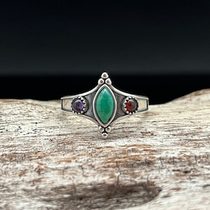 May include: A silver ring with a green gemstone in the center and two smaller gemstones on either side. The smaller gemstones are purple and red.