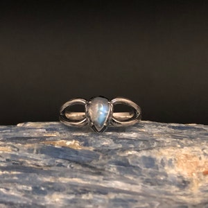 May include: A silver ring featuring a teardrop-shaped moonstone set in the centre. The ring has a delicate design with two curved bands that meet at the stone.