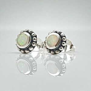 May include: A pair of stud earrings featuring round, iridescent white opals set in oxidized sterling silver. Each earring is surrounded by a decorative beaded border, creating a textured, circular frame around the central gemstone.