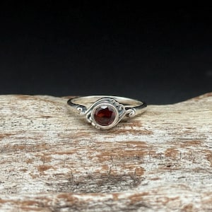 May include: A silver ring with a round, deep red gemstone at the center. The ring has a decorative design around the gemstone and on the band. The ring is displayed on a piece of wood.