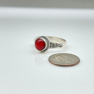 May include: A silver ring with a round, faceted red stone. The ring has a decorative rope-like border around the stone and scrollwork on the band. A US dime is next to the ring for size comparison.