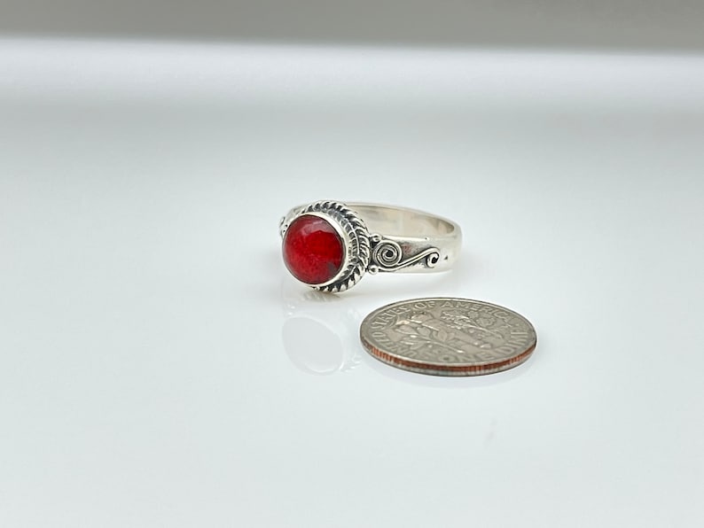 May include: A silver ring with a round, faceted red stone. The ring has a decorative rope-like border around the stone and scrollwork on the band. A US dime is next to the ring for size comparison.