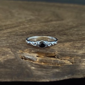 May include: A silver ring with a small, round, black gemstone at the center. The ring has a delicate, ornate design with swirling patterns on the band. The ring is displayed on a wooden surface.