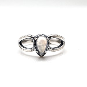 May include: A silver ring featuring a pear-shaped white gemstone set in the centre. The ring has a distinctive design with two curved bands that meet at the gemstone.