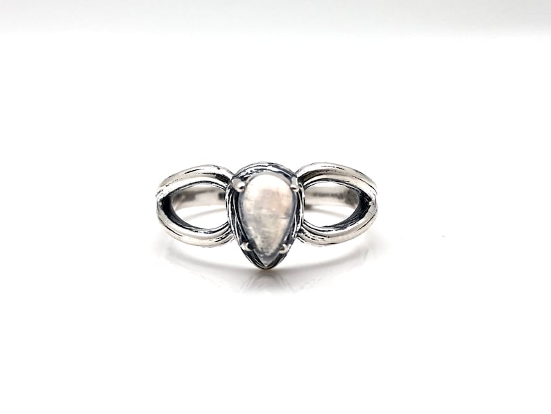 May include: A silver ring featuring a pear-shaped white gemstone set in the centre. The ring has a distinctive design with two curved bands that meet at the gemstone.