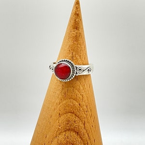 May include: A silver ring with a round, vibrant red stone. The ring has a decorative rope-like border around the stone and scrollwork on the band. The ring is displayed on a wooden cone-shaped stand against a white background.