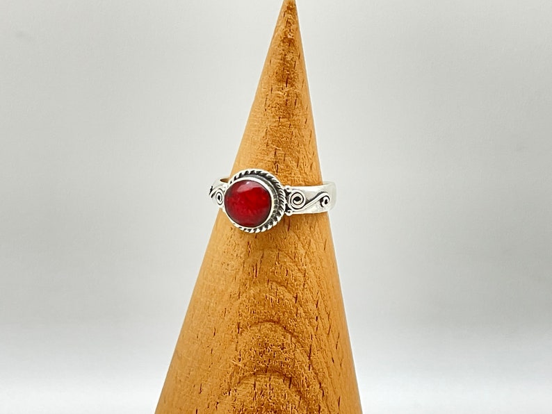 May include: A silver ring with a round, vibrant red stone. The ring has a decorative rope-like border around the stone and scrollwork on the band. The ring is displayed on a wooden cone-shaped stand against a white background.