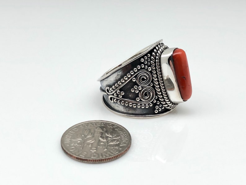 May include: A silver ring with an orange stone set in a decorative band. The band is detailed with a rope design and a swirl pattern.