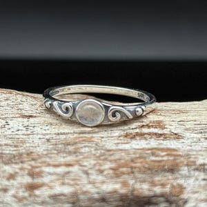 Puede incluir: Un anillo de plata con una piedra pequeña, redonda y de color claro en el centro. El anillo presenta adornos de volutas en la banda. El anillo se muestra sobre un trozo de madera.