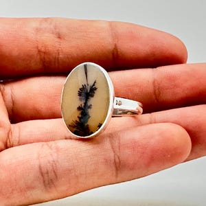 K&ouml;nnte beinhalten: Silberring mit einem ovalen Dendritachat-Stein. Der Stein hat einen cremefarbenen Hintergrund mit schwarzen, baumartigen Einschl&uuml;ssen. Der Ring ist in einem polierten Silberband gefasst, ein einzigartiges Schmuckst&uuml;ck.