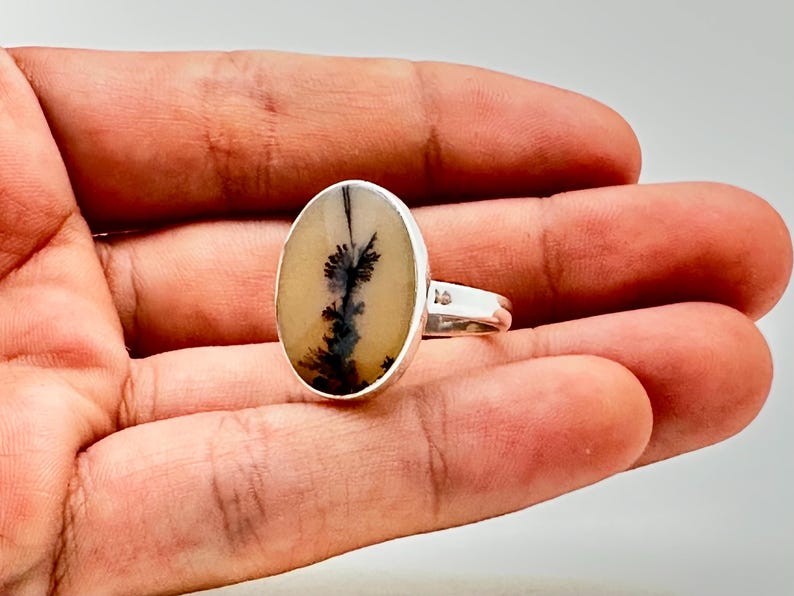 K&ouml;nnte beinhalten: Silberring mit einem ovalen Dendritachat-Stein. Der Stein hat einen cremefarbenen Hintergrund mit schwarzen, baumartigen Einschl&uuml;ssen. Der Ring ist in einem polierten Silberband gefasst, ein einzigartiges Schmuckst&uuml;ck.