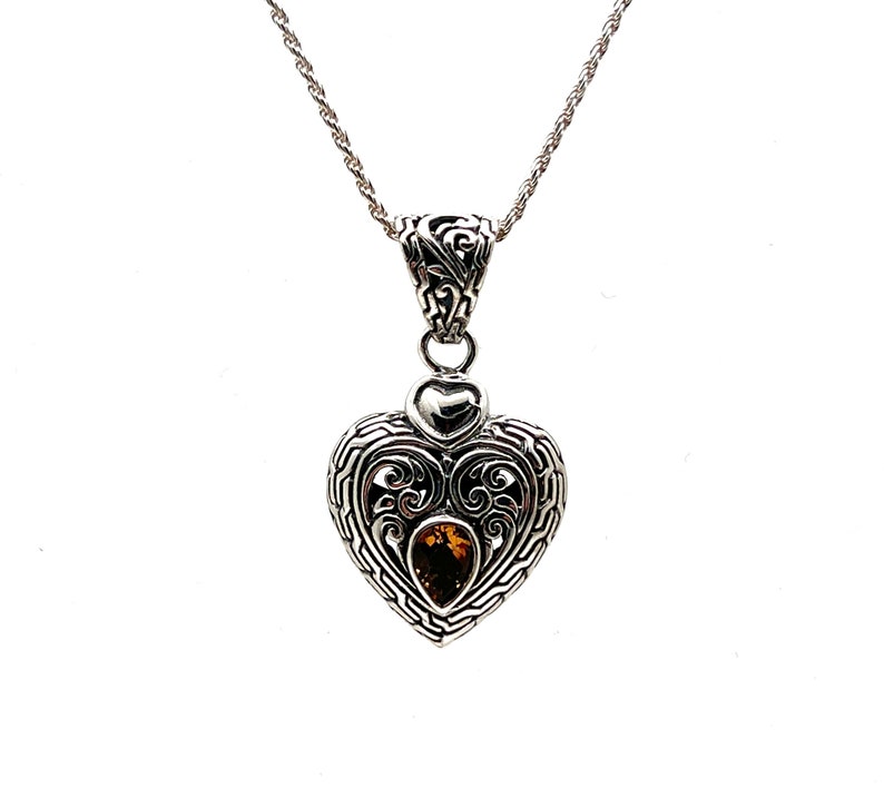 Puede incluir: Un collar con un colgante en forma de coraz&oacute;n de plata con un intrincado dise&ntilde;o de filigrana y una piedra preciosa marr&oacute;n en el centro.