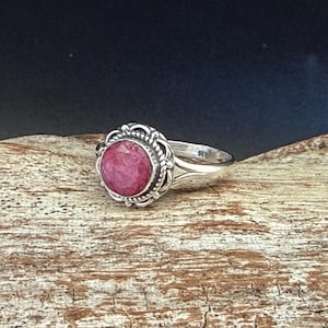 May include: A sterling silver ring with a large, round, faceted ruby gemstone. The ruby is a vibrant pinkish-red color, set in a decorative, ornate bezel. The ring sits on a textured, light brown wooden surface.