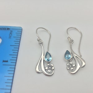 May include: A pair of silver dangle earrings featuring a teardrop shaped blue topaz gemstone and a flower design.