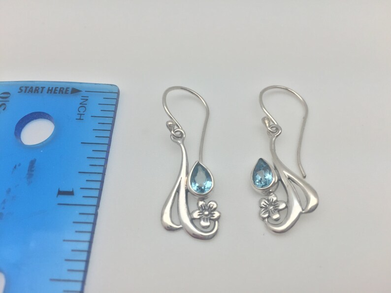 May include: A pair of silver dangle earrings featuring a teardrop shaped blue topaz gemstone and a flower design.