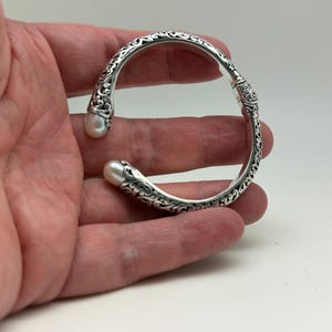 May include: Silver-toned cuff bracelet with ornate scrollwork and two white pearls at the ends. The bracelet has an open design and is held in a hand. The bracelet is a piece of jewelry.