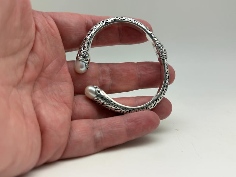 May include: Silver-toned cuff bracelet with ornate scrollwork and two white pearls at the ends. The bracelet has an open design and is held in a hand. The bracelet is a piece of jewelry.
