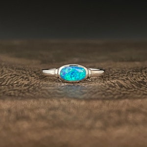 May include: A silver ring with an oval, blue-green opal stone. The ring's band is thin and polished, and the opal has a vibrant, iridescent quality. The ring is set against a dark, textured background.