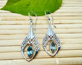 May include: A pair of silver drop earrings featuring an openwork teardrop frame with intricate scrollwork patterns. Each earring suspends a single marquise-cut blue gemstone in the center, attached to a curved silver ear wire for secure hanging.