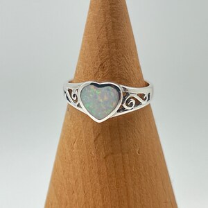 May include: A silver ring with a heart-shaped opal gemstone. The ring has a delicate, scroll-like design.