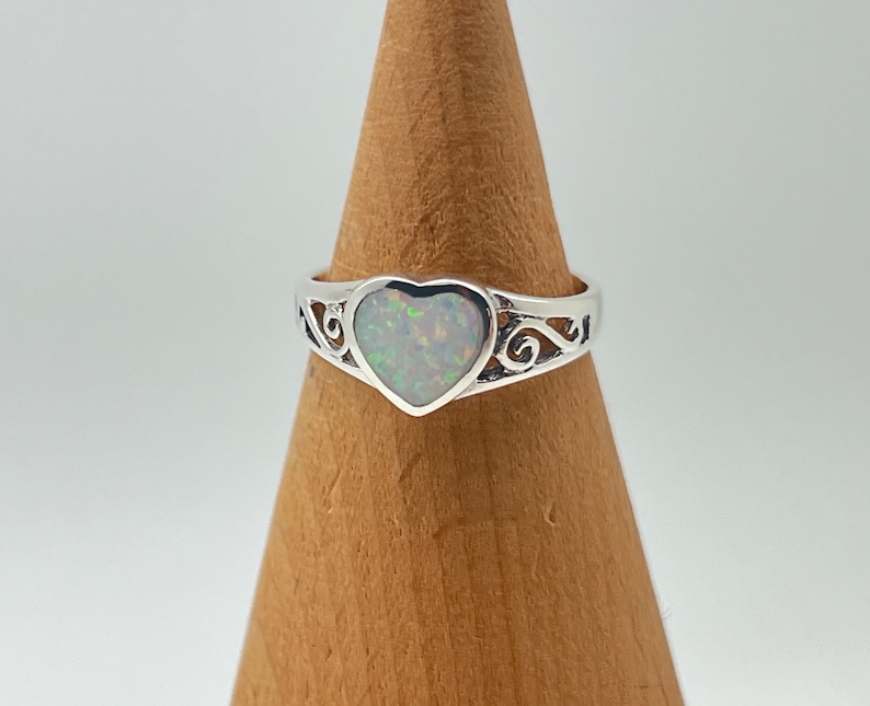 May include: A silver ring with a heart-shaped opal gemstone. The ring has a delicate, scroll-like design.