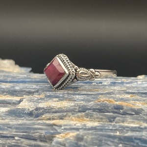 May include: A silver ring featuring a square ruby gemstone set in an ornate bezel. The ring has a textured band and intricate detailing.