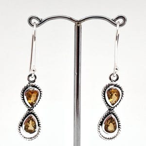 May include: A pair of silver drop earrings featuring two teardrop-shaped citrine gemstones each, set in a twisted rope-style metal frame. The earrings hang from simple silver hooks and display a warm yellow-orange hue in the faceted stones.
