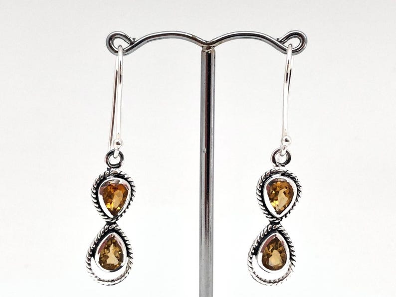 May include: A pair of silver drop earrings featuring two teardrop-shaped citrine gemstones each, set in a twisted rope-style metal frame. The earrings hang from simple silver hooks and display a warm yellow-orange hue in the faceted stones.