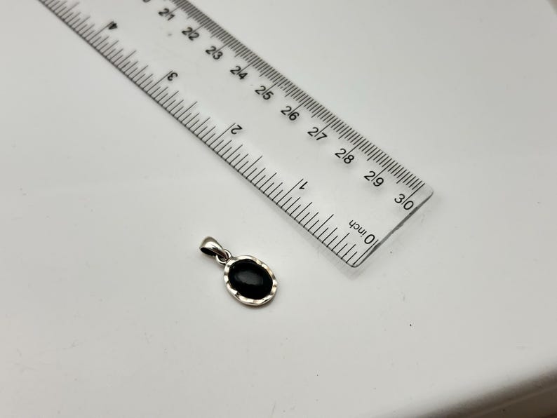 Puede incluir: Un colgante de plata con una piedra negra ovalada, junto a una regla. El colgante mide aproximadamente 2.5 cm de largo. La regla est&aacute; marcada en pulgadas.