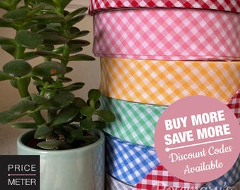 May include: A stack of fabric ribbons featuring a gingham check pattern in red, pink, yellow, green, blue, and gray. A pink circular graphic overlay displays the text BUY MORE SAVE MORE Discount Codes Available. A small green plant is visible.